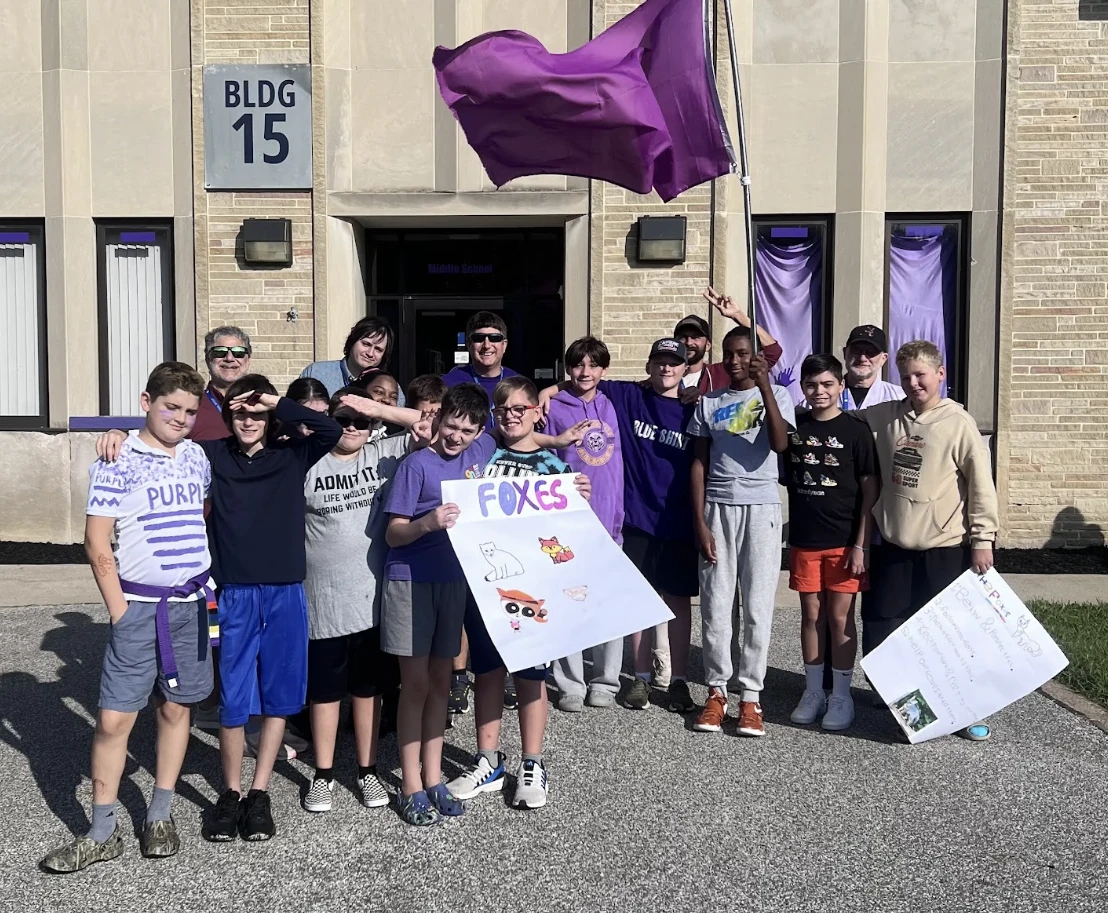 Students and staff with a purple flag and a “Foxes” sign outside Building 15 on campus
