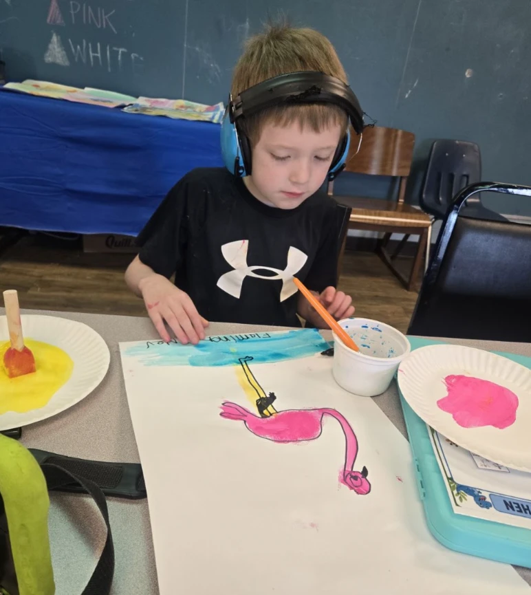 A student wearing ear protection paints a pink flamingo at an art table