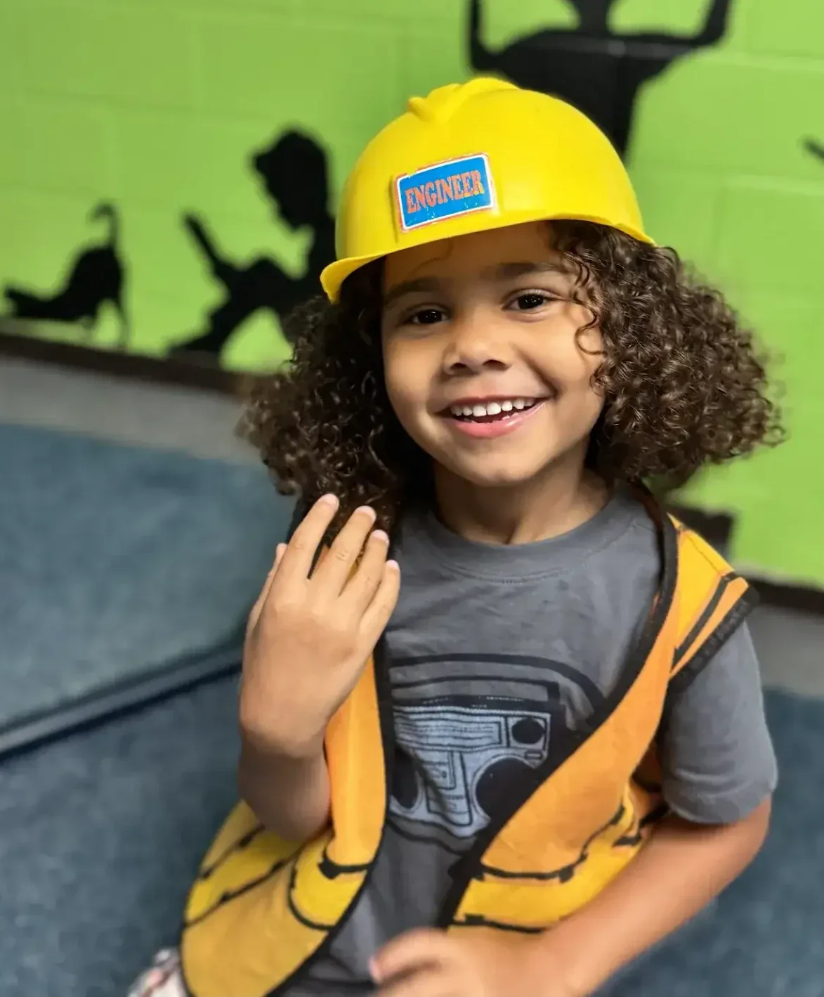A smiling student wearing a yellow toy hard hat labeled “Engineer” and an orange safety vest
