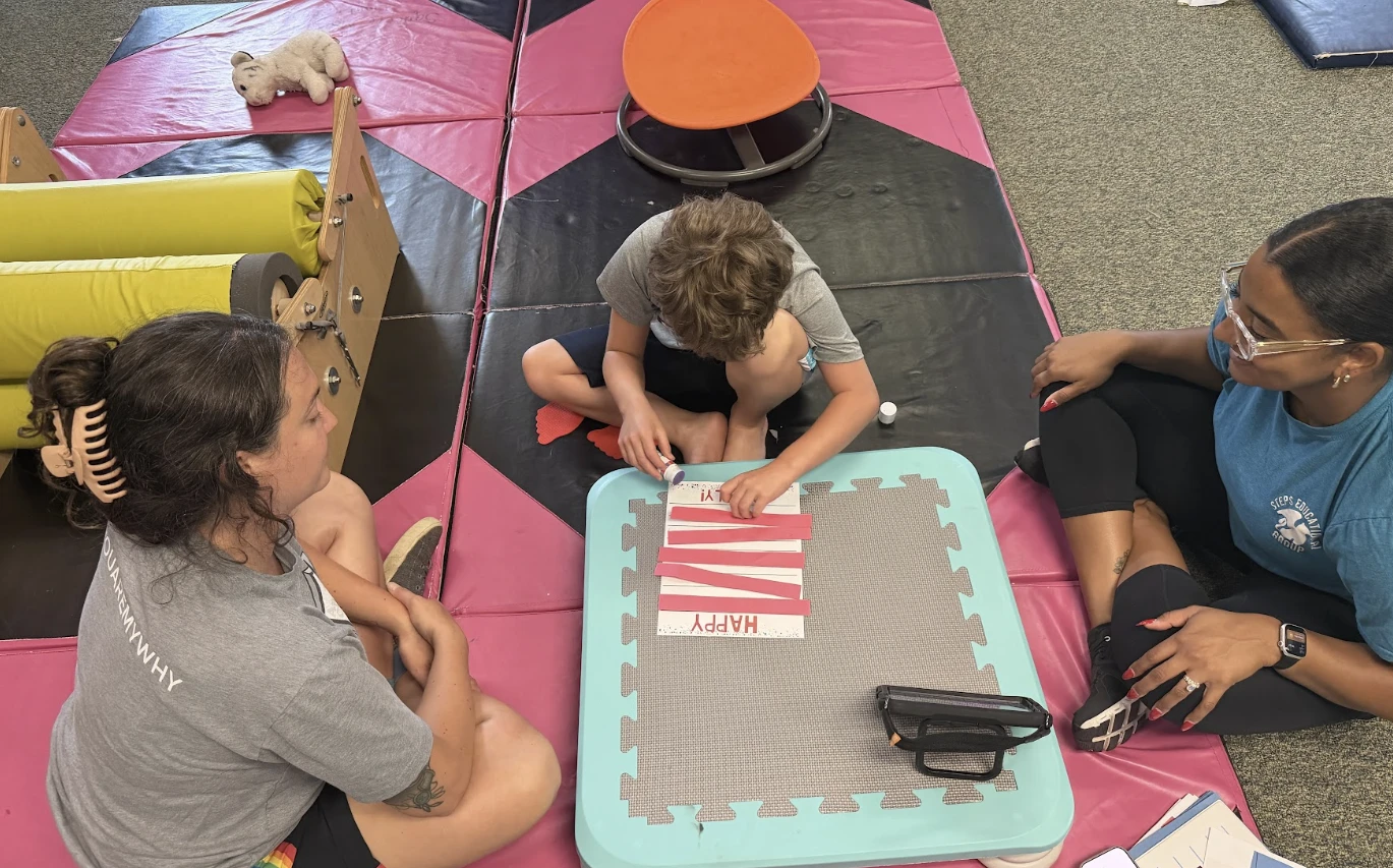 A student works on a craft at a low table in the sensory room with two staff members