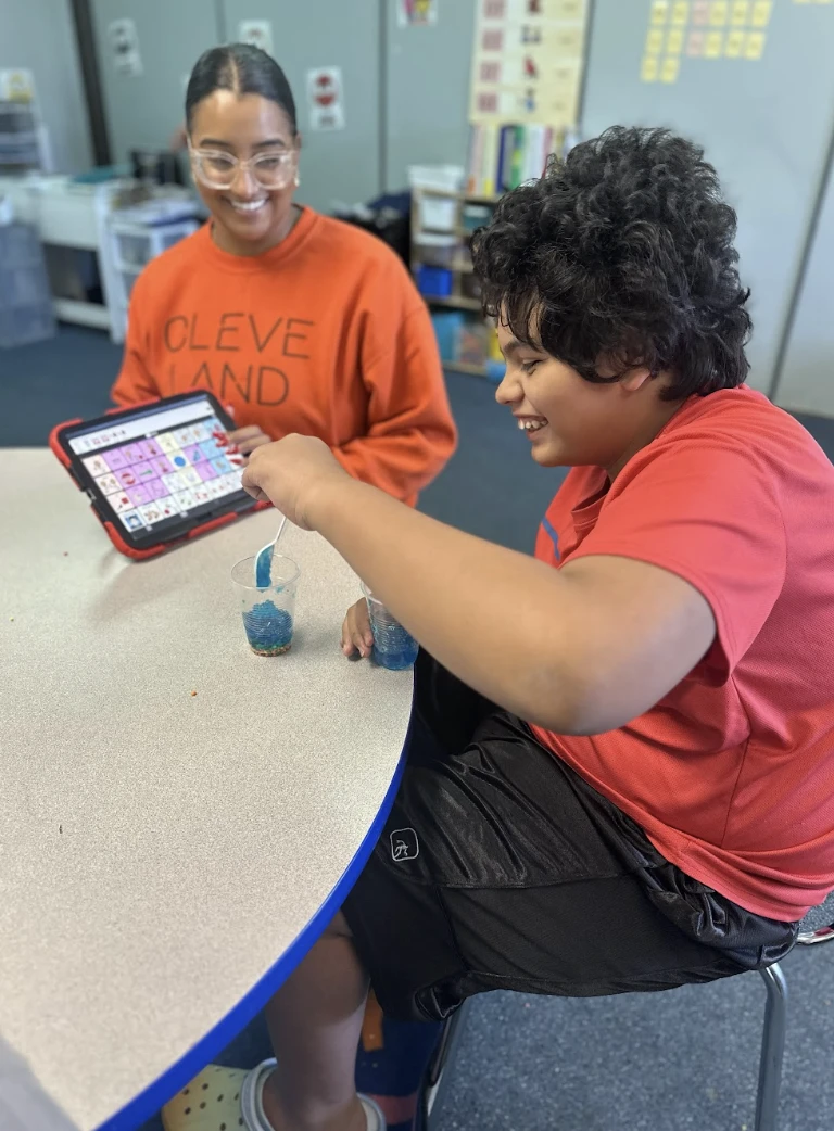 A smiling student works on a classroom activity while a teacher holds up the student’s communication device